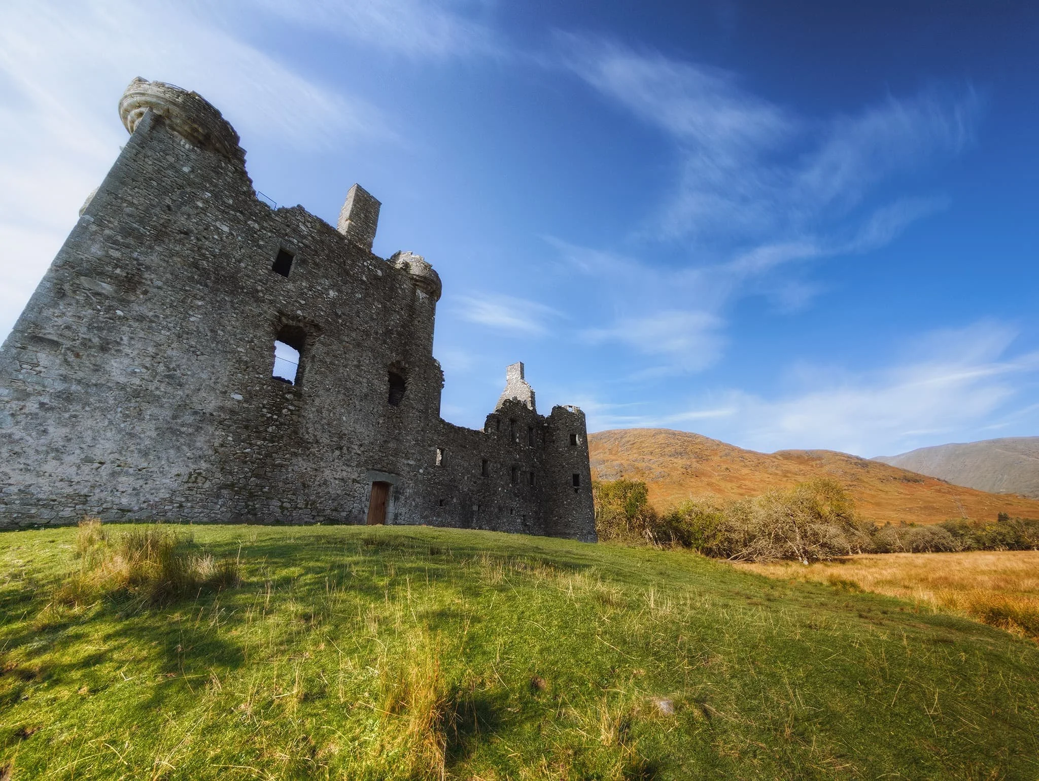 An ultra-wide perspective of the front of Kilchurn Castle, with the slopes of Ben Cruachan in the distance and some beautiful wispy clouds above.