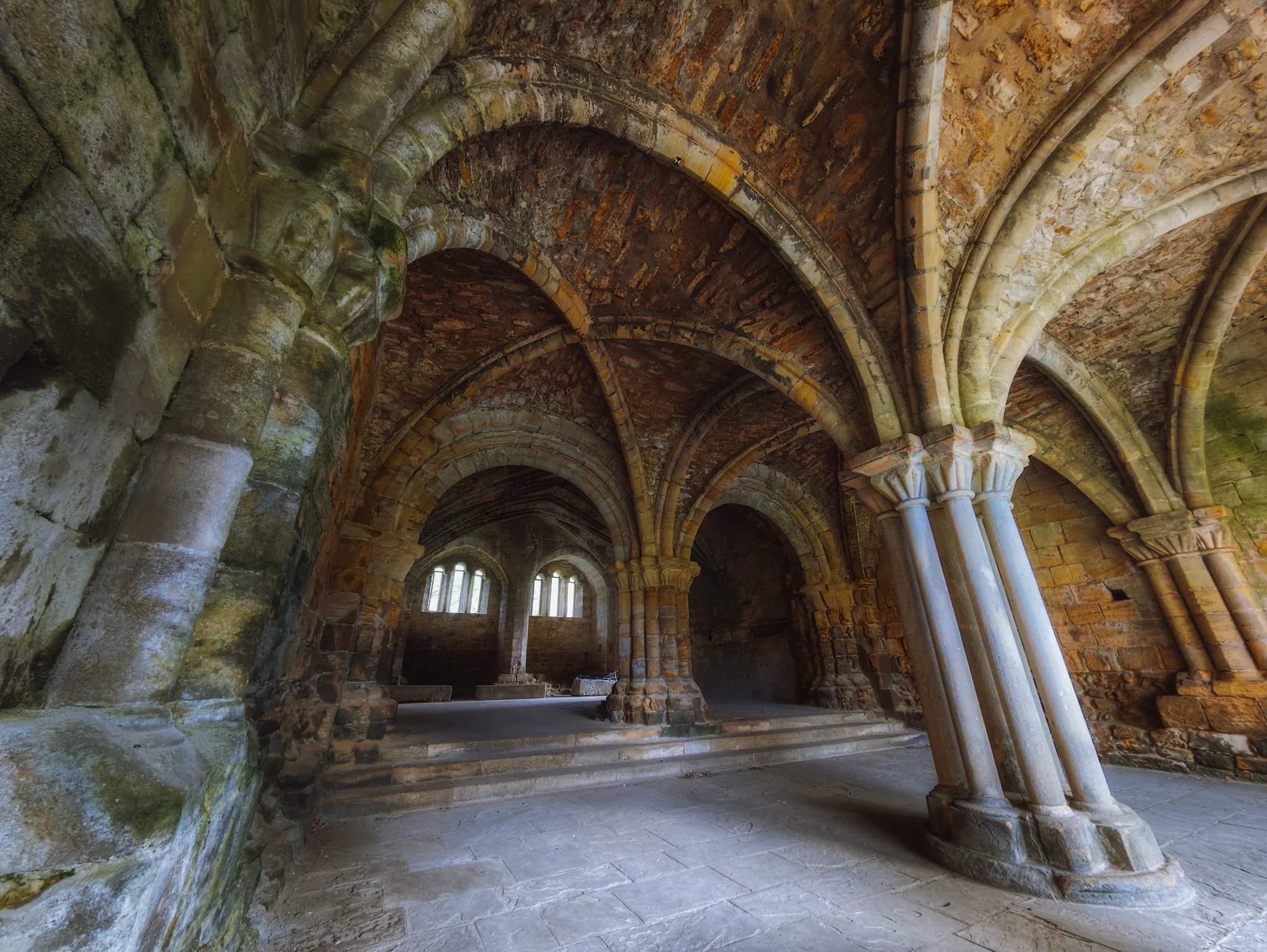 Looking into the Chapter House of Kirkstall Abbey. It features a textbook example of Transitional Architecture, marking the shift from the heavy Norman style to the lighter Gothic era. You can see this mix in the combination of rounded window arches with the newer structural ribbed vaulting overhead.