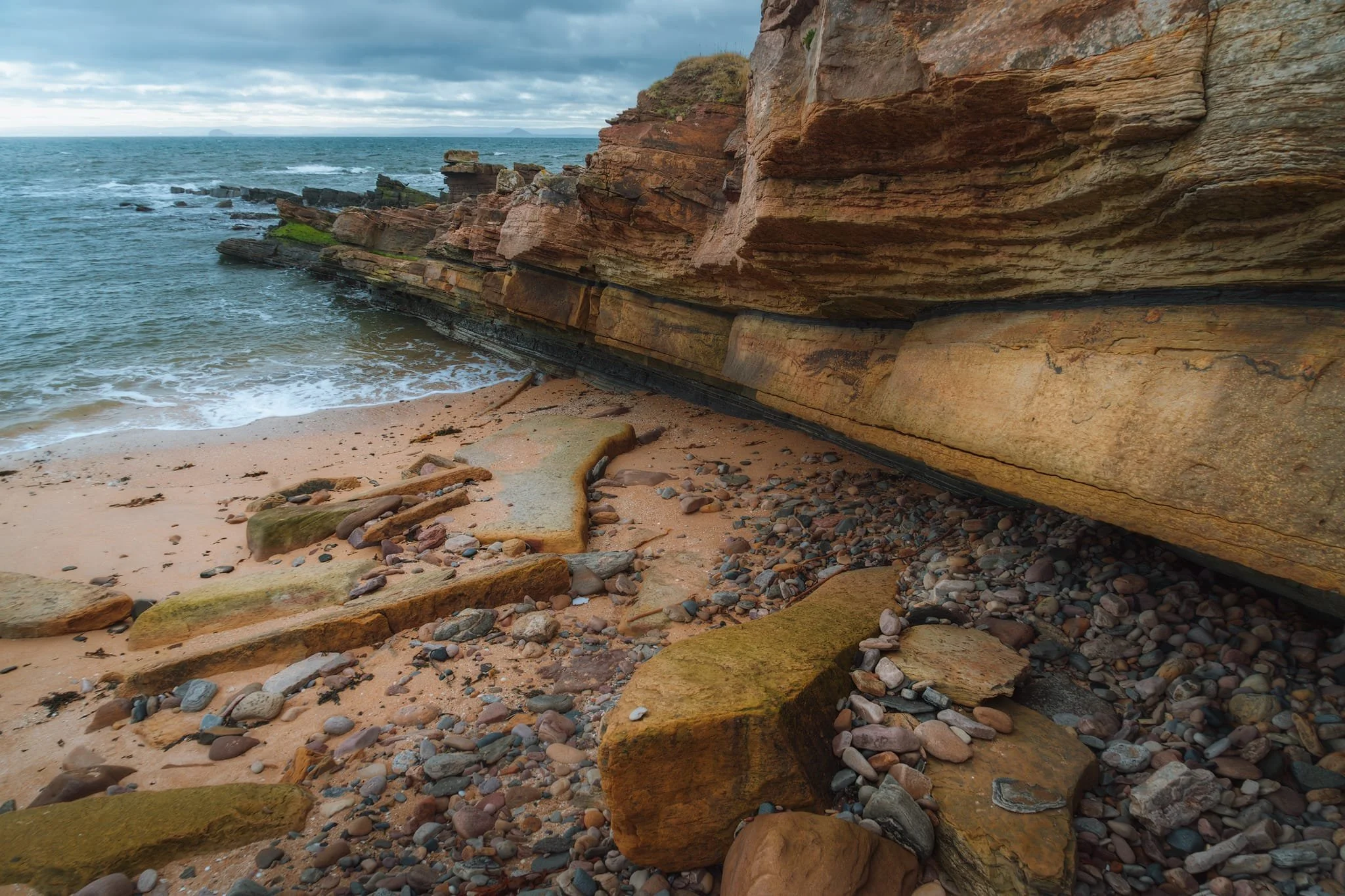 Below the playground and Anstruther Wester’s golfing area, one can play around with the crazy rock formations at Billow Ness. The cliffs and boulders are primarily composed of sandstone; the layering indicates these rocks were deposited in horizontal strata, like an ancient river or sea. The reddish-brown colouring hints at the presence of iron oxides. In the distance, across the Firth of Forth, you can just about make out two distinct “bumps” on the horizon. The one on the left is Bass Rock, and on the right is Berwick Law.