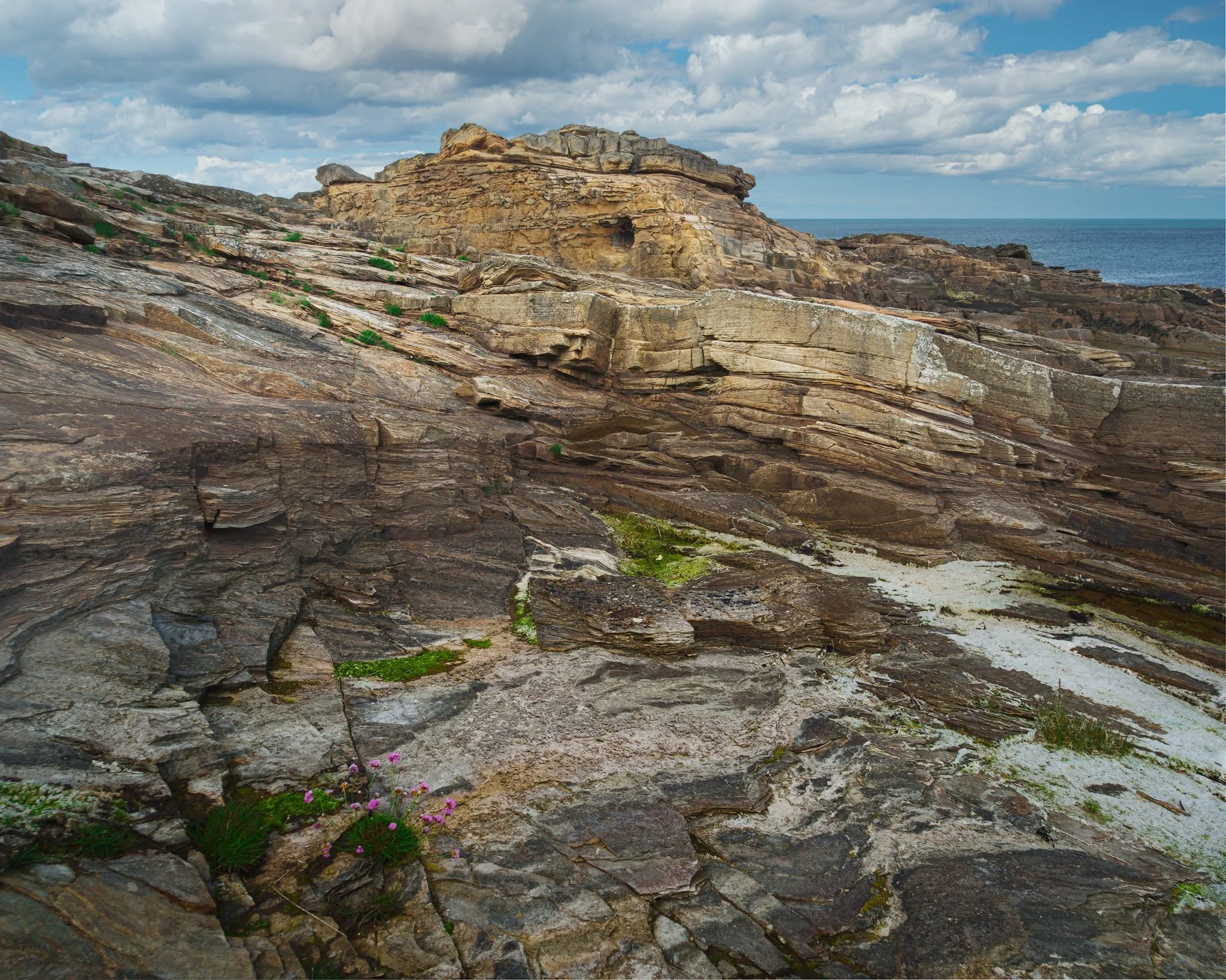 A fantastical mish-mash of geology here. In the foreground, smooth, low-angle siltstones/mudstones formed from old overbank (floodplain) mud settling out when the river crept over its banks. Modern tides, salt spray and lichens have bleached and smoothed those laminations, making them look chalky. The subtle, sharp break (you can just make it out sloping away) marks where a later channel scoured into those muds. The big yellow-buff blocks are the Rumbling Kern channel sandstones: coarse, cross-bedded braid-plain sand dumped by fast currents.